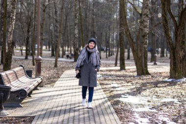 Sıcak kıyafetler ve beyaz spor ayakkabılar giymiş bir kadın baharın başında güneşli bir şehir parkında yürür..