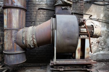 Terk edilmiş bir metal işleme tesisinde eski paslı bir havalandırma ünitesi. Arka planda siyah dumanlı tuğla duvar