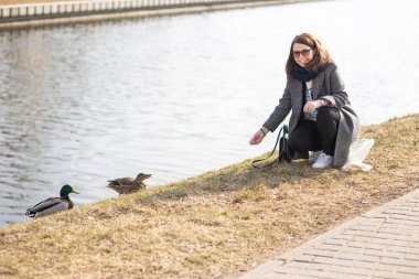 Güneş gözlüklü, şık elbiseli bir kadın gülüyor ve güneşli bir bahar parkında nehir kenarındaki ördekleri besliyor..