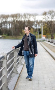 Gözlüklü bir adam nehir kenarında şehir parkında yürüyor. Adam mavi bir pelerin ve çizgili bir eşarp giyiyor.