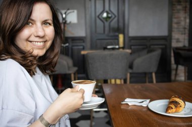 Bir kadın, boş bir kafede elinde bir fincan kapuçino ile oturmuş gülümsüyor. Meşe ağacının üstünde kruvasan olan bir tabak var.