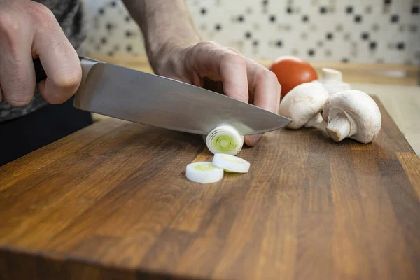 The chef slices leeks on an oak board. There are mushrooms and a tomato on the board next to it.