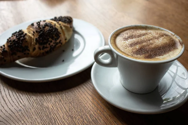 There is a cup of cappuccino on an oak table. Blurred croissant on a plate on background