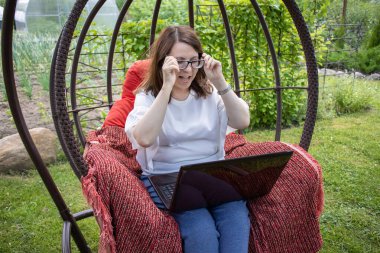 Uzun saçlı bir kadın yeşil bir yaz bahçesinde yuva sandalyesinde otururken dizüstü bilgisayar ekranına bakarken seviniyor.