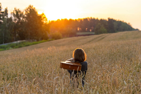 A girl with long hair with an old acoustic guitar stands in a wheat field lit by the rays of the setting sun and admires the sunset