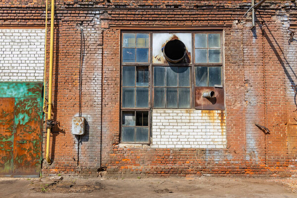 A large old dilapidated house of red and white bricks with a large dilapidated window and a multi-colored metal door. Old dirty abandoned factory building.