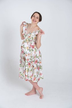 Beyaz gri arka planda, mutlu hamilelik kavramı üzerine yaz elbisesi giymiş neşeli hamile kadının stüdyo portresi.