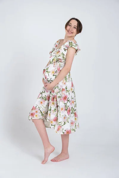 Studioporträt einer fröhlichen jungen Schwangeren im Sommerkleid auf weißgrauem Hintergrund, glückliches Schwangerschaftskonzept — Stockfoto