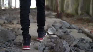 Bir genç elinde baltayla harabelerde yürüyor.