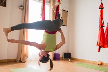 Kırmızı bir yoga hamağında ters dönmüş bir kadın, kapalı bir stüdyoda zorlu bir esneklik pozu veriyor.