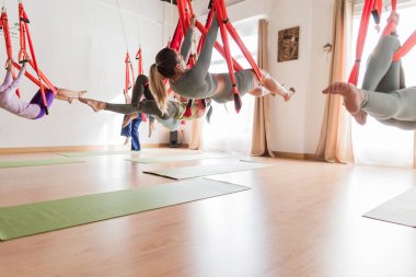 Çok kültürlü bir gruptaki kadınlar, vinyasa kursu sırasında kumaş hamaklarda asılı halde havadan yoga egzersizleri yapıyorlar.