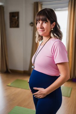 Hamile kadın yoga stüdyosunda ayakta, gülümsüyor ve karnına dokunuyor. Doğum öncesi sağlık ve anneliğe hazırlanıyor.