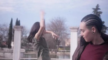 Saçları örgülü şık bir beatboksör arka planda serbest dans eden bir kadın. İki genç sanatçı açık havada güneşli bir günün tadını çıkarıyor.