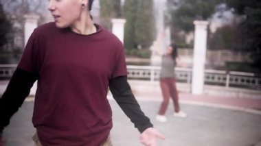 Saçları örgülü genç bir kadın ön planda boks yapıyor. Başka bir kadın parkta serbest dans edip hip hop yapıyor.