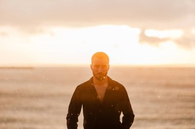 Gün batımında denizin kenarında duran genç adam, yoğun, şehvetli bir ifadeyle düşünüyor, yalnızlığı ve ilhamı somutlaştırıyor.