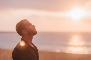 Gözleri kapalı, başı eğik, plajda günbatımının sıcaklığının ve huzurunun tadını çıkaran genç bir adamın profil görüntüsü.