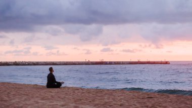 Genç adam kumlu sahilde meditasyon pozunda oturur, sakin günbatımında ya da gün doğumunda okyanus ve ufku düşünür.