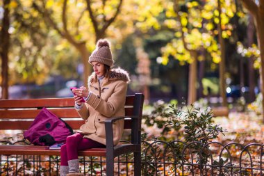 Kışın giyinmiş bir kadın, sonbahar ağaçları ve yapraklarla çevrili bir bankta otururken akıllı telefonuna odaklanmış.