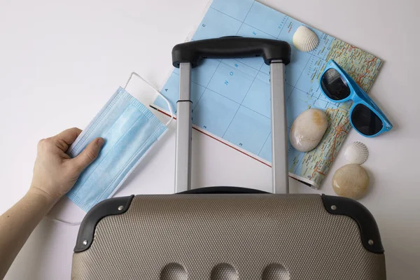 Travel during the time of COVID-19. Medical mask, suitcase, sun glasses, map, seashells on the white background. Vacation, holidays in corona times. — Stock Photo, Image