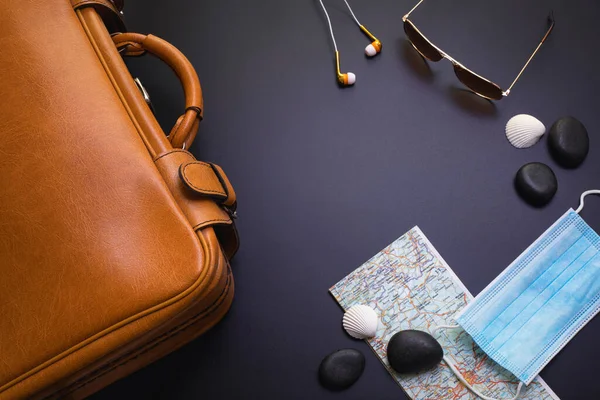 Reisen während der Zeit von COVID-19. Medizinische Maske, Koffer, Sonnenbrille, Karte, Muscheln, Kopfhörer auf schwarzem Hintergrund. Urlaub, Urlaub in Corona-Zeiten. — Stockfoto