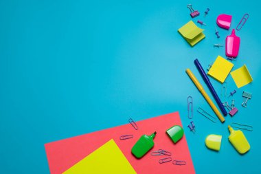 Mavi arkaplanda metin için boş alanı olan kırtasiye ögeleri. Yaratıcı, renkli arka plan, okul veya ofis malzemeleri. Flatlay ile kopyalama alanı, üst görünüm. Kalemler, ataçlar, yapışkan notlar..
