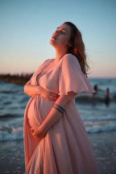 Young pregnant woman with a beautiful sea view on the background. Happy and calm pregnant woman with long hair and pink dress standig on the beach. Maternity. — Stock Photo, Image