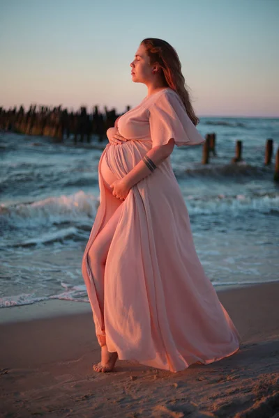 Jeune femme enceinte avec une belle vue sur la mer sur le fond. Joyeux et calme femme enceinte aux cheveux longs et robe rose standig sur la plage. Maternité. — Photo