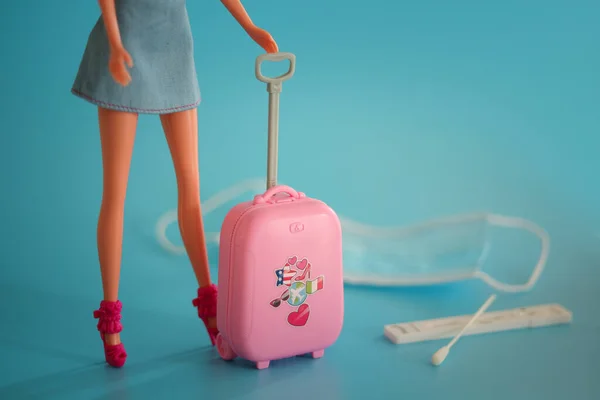 Doll with suitcase. Travel and flights during the time of COVID-19. Doll, medical mask, coronavirus antigentest on the blue background. Vacation, holidays in corona times. Colorful design. — Stock Photo, Image