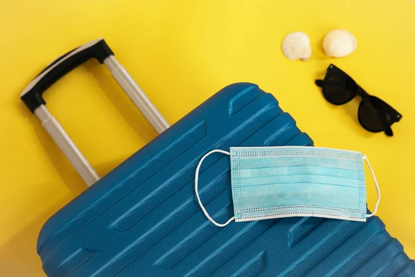 Travel during the time of COVID-19. Medical mask, suitcase, sun glasses, seashells on the yellow background. Vacation, holidays in corona times. — Stock Photo, Image