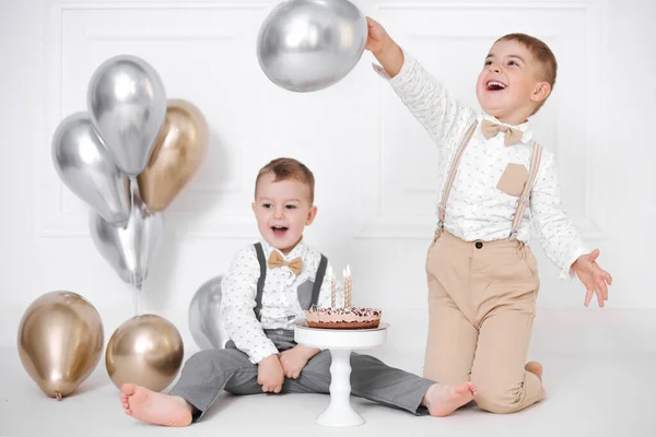 Två pojkar firar födelsedag, barn har en B-dagars fest. Födelsedagstårta med ljus och ballonger. Glada barn, fest, vit minimalistisk interiör. — Stockfoto