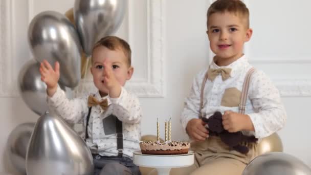 Deux garçons fêtent leur anniversaire, les enfants applaudissent. Gâteau d'anniversaire avec bougies et ballons. Enfants heureux, célébration, intérieur minimaliste blanc. — Video