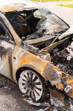 Yanmış araba enkazı kömürleşmiş metal ve küllerle büyük hasar gösteriyor. Araç sanayi bölgesinde yol kenarında duruyor, yakın zamanda çıkan bir yangının belirtilerini gösteriyor..