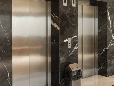 Lüks bir lobide koyu mermer duvarlara karşı iki parlak paslanmaz çelik asansör görülebilir. Yakınlarda bir çöp tenekesi duruyor, modern estetiğe katkı sağlıyor..