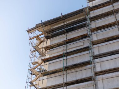 İşçiler bir kentteki yüksek bir binayı yenilemek için iskeleyi kullanıyorlar. Güçlü güneş ışığı yapıyı ve devam eden inşaat faaliyetlerini vurguluyor.