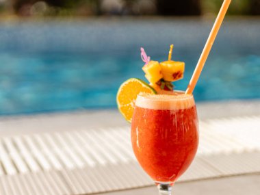 Portakal ve ananasla süslenmiş renkli bir kokteyl sıcak güneşin altında ışıldayan bir havuz kenarında bir masada oturur. Rahatlamak ve eğlenmek için ideal..