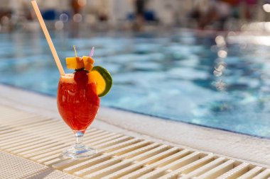 Taze meyve ve renkli bir şemsiyeyle süslenmiş ferahlatıcı tropikal bir kokteyl havuzun kenarında oturur. Güneş ışığı suyu yansıtır ve canlı bir atmosfer yaratır..