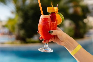 Bir el köpüklü bir havuzun yanında canlı bir tropikal kokteyl tutar, parlak renkler ve süslemeler sergiler, güneşli bir gün ve keyif için mükemmeldir. Tüm kapsayıcı kavramlar