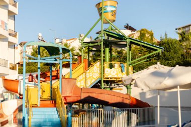 Canlı bir su kaydırağı kompleksi, birden fazla slayt ve su sıçratmak için büyük bir kova içeren bir yaz otelinde duruyor. Palmiye ağaçları ve açık gökyüzü neşe dolu atmosfere katar..