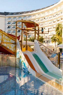 Konuklar güneşli bir günde otel havuzunda eğleniyorlar. Ana ilgi odağı canlı bir su kaydırağıydı. Arkaplanda modern mimari ve palmiye ağaçları yer alıyor..
