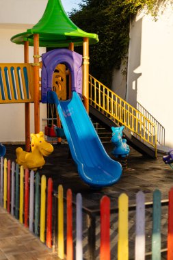 Canlı bir oyun parkında mavi bir slayt, oyuncu hayvan figürleri ve renkli eskrim figürleri bulunur. Güneşli ortam çocukları eğlenmeye ve oynamaya davet ediyor..