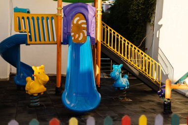 Canlı bir oyun alanı sahnesi. Mavi bir slayt, sarı hayvan yay binicileri ve parlak yapılar. Bu güvenli oyun alanı çocukların keşfetmesi ve eğlenmesi için mükemmel..