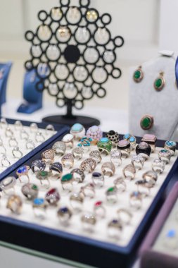 Bir kuyumcunun masasında renkli bir dizi yüzük ve küpe sergileniyor. Parlak mücevherler ve karmaşık tasarımlar ile dolu bir pazar gününde alıcıları cezbediyor..