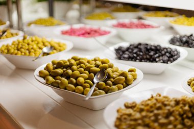 Canlı bir zeytin seçimi bir gıda pazarında sunuluyor, beyaz kaselerde yeşil, siyah ve pembe çeşitleri sergileniyor. Misafirler eşsiz lezzet ve dokuları tatmaktan hoşlanırlar..