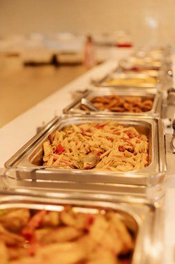Farklı sos ve malzemelerle dolu makarna tabakları uzun bir masanın üzerindeki metal tepsilerde serilidir. Bu büfe, misafirleri kendilerine hizmet etmeleri için davet ediyor..