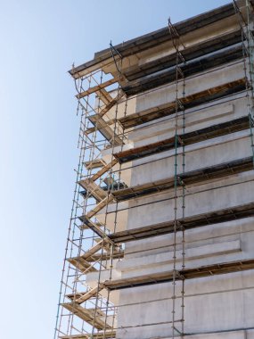 İşçiler yenileme görevlerini yerine getirirken, iskele yüksek bir binayı çevrelemektedir. Mavi gökyüzü şehirdeki inşaat alanı için bir zemin sağlıyor.