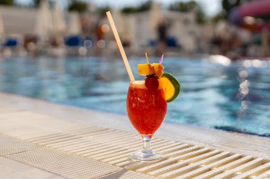 Parlak bir kokteyl havuzun kenarında durur. Meyve süslemeleri ve pipetleri var. İnsanlar açık mavi gökyüzünün altında güneşlenir ve yüzer..