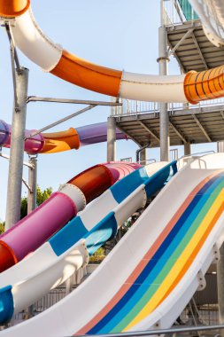 Çocuklar ve yetişkinler renkli su kaydıraklarıyla dolu bir su parkında günün tadını çıkarıyorlar. İnsanlar eğlence ve heyecan için slaytların yanında toplanırken güneş ışıl ışıl parlıyor.