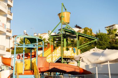 Çocuklar su kaydırağının yanında birden fazla kaydırak ve aşağı dökülen büyük bir su kovası ile oynuyorlar. Yapı güneşli bir gökyüzünün altında ağaçlar ve binalarla çevrili..