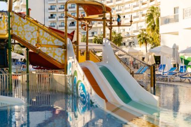 Çocuklar tatil köyü havuzundaki su kaydırağının tadını çıkarabilirler. Aileler şemsiyelerin altındaki şezlonglarda dinlenirler. Bu sahne bir su parkındaki yaz aktivitelerini gösteriyor..