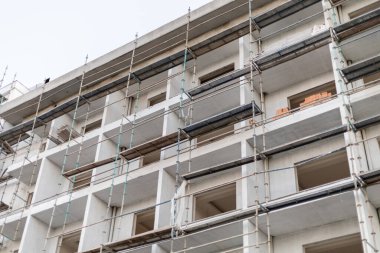 İşçiler çok katlı bir binada inşaat işleriyle meşguller. İskeleler yapıyı çevrelemiş, gelişimin çeşitli aşamalarını göstermektedir. Araçlar ve malzemeler görünür..